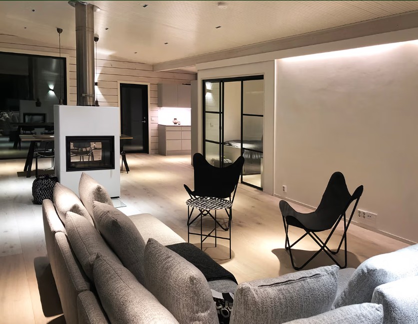Moderni olohuone, jossa harmaa sohva, mustat tuolit, takka ja taustalla avokeittiö – tunnelman ja tyylin muotoiluee LedStore.fi:n laadukas led-valaistusratkaisu.